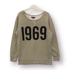 May include: A light brown sweatshirt with long sleeves and a crew neck. The sweatshirt has a large black print of the number "1969" on the front.