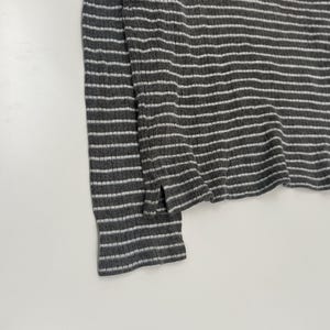 May include: A long-sleeved shirt featuring a dark gray and white horizontal striped pattern. The garment is crafted from a ribbed knit fabric and includes a small side slit at the hemline. A small, rectangular logo patch is visible near the neckline.