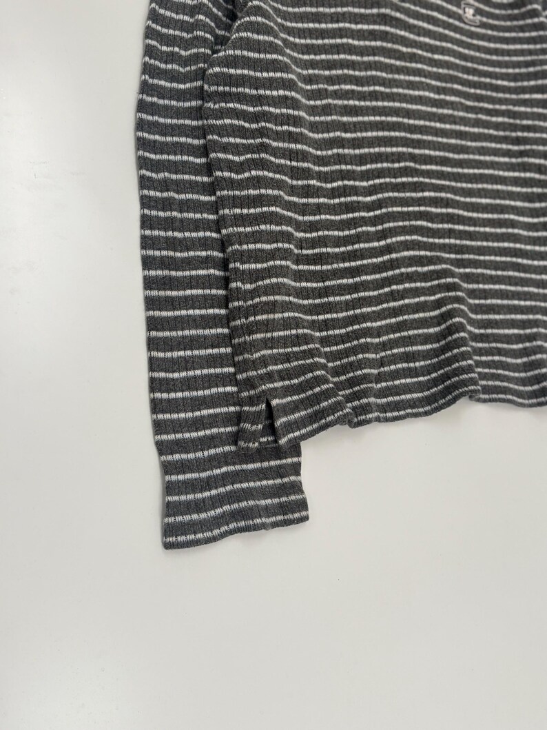 May include: A long-sleeved shirt featuring a dark gray and white horizontal striped pattern. The garment is crafted from a ribbed knit fabric and includes a small side slit at the hemline. A small, rectangular logo patch is visible near the neckline.