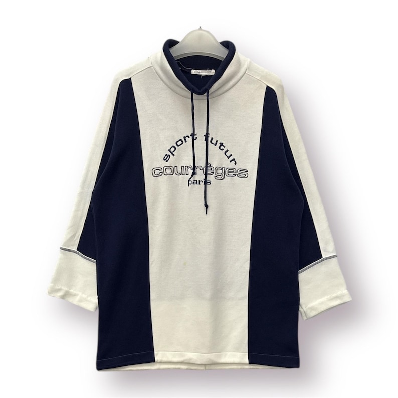 Puede incluir: Una sudadera crema y azul marino con el texto "sport futur courr&egrave;ges paris". La sudadera tiene un cuello alto con cord&oacute;n y mangas tres cuartos. El dise&ntilde;o presenta paneles azul marino en los laterales.