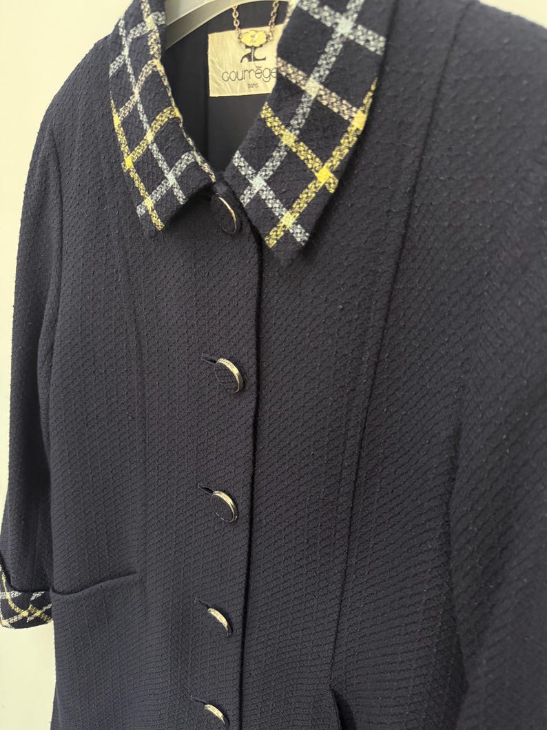 May include: A navy blue jacket with a textured fabric. The jacket features a plaid collar with yellow and white accents. The jacket has four silver buttons and a small pocket. The label reads "courr&egrave;ges paris".