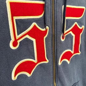 May include: Close-up of a navy blue zip-up hoodie with the number "55" prominently displayed on the front. The number is red with a cream-colored border and yellow accents. The hoodie has a drawstring and a brass zipper.