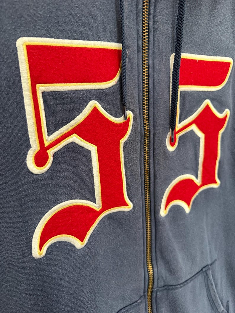 May include: Close-up of a navy blue zip-up hoodie with the number "55" prominently displayed on the front. The number is red with a cream-colored border and yellow accents. The hoodie has a drawstring and a brass zipper.