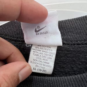 Puede incluir: Primer plano de una etiqueta blanca con el logotipo de Nike y texto. La etiqueta incluye instrucciones de lavado: 50% algod&oacute;n, lavar a m&aacute;quina en fr&iacute;o, secar en secadora a baja temperatura, planchar en fr&iacute;o, lej&iacute;a sin cloro seg&uacute;n sea necesario. La talla es XL (18-20).