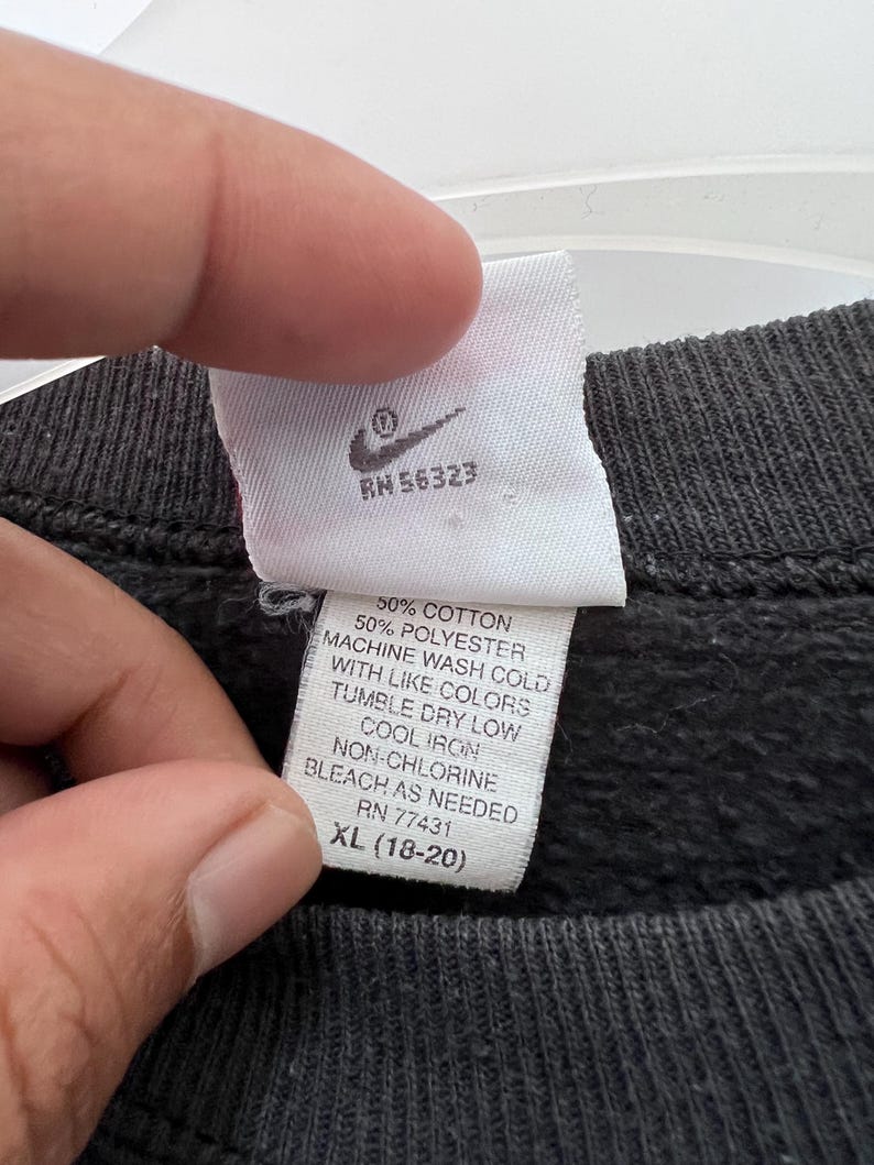 Puede incluir: Primer plano de una etiqueta blanca con el logotipo de Nike y texto. La etiqueta incluye instrucciones de lavado: 50% algod&oacute;n, lavar a m&aacute;quina en fr&iacute;o, secar en secadora a baja temperatura, planchar en fr&iacute;o, lej&iacute;a sin cloro seg&uacute;n sea necesario. La talla es XL (18-20).