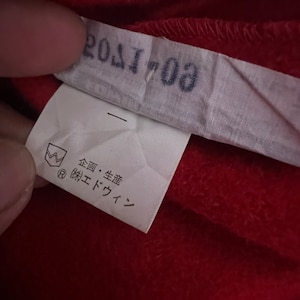 May include: A white tag with black text that reads "201703". The tag also has a small black and white symbol with a crown and the letter "R" inside a circle. Below the symbol is more black text that reads "企画・生産 (株) エドウイン".