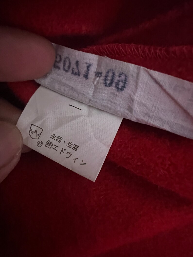May include: A white tag with black text that reads "201703". The tag also has a small black and white symbol with a crown and the letter "R" inside a circle. Below the symbol is more black text that reads "企画・生産 (株) エドウイン".