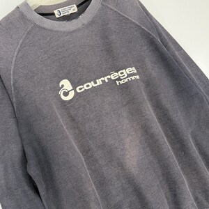 May include: A gray crew neck sweatshirt with the text "courr&egrave;ges homme" in white. The sweatshirt has a ribbed texture and a faded appearance. The brand label is visible at the neckline.