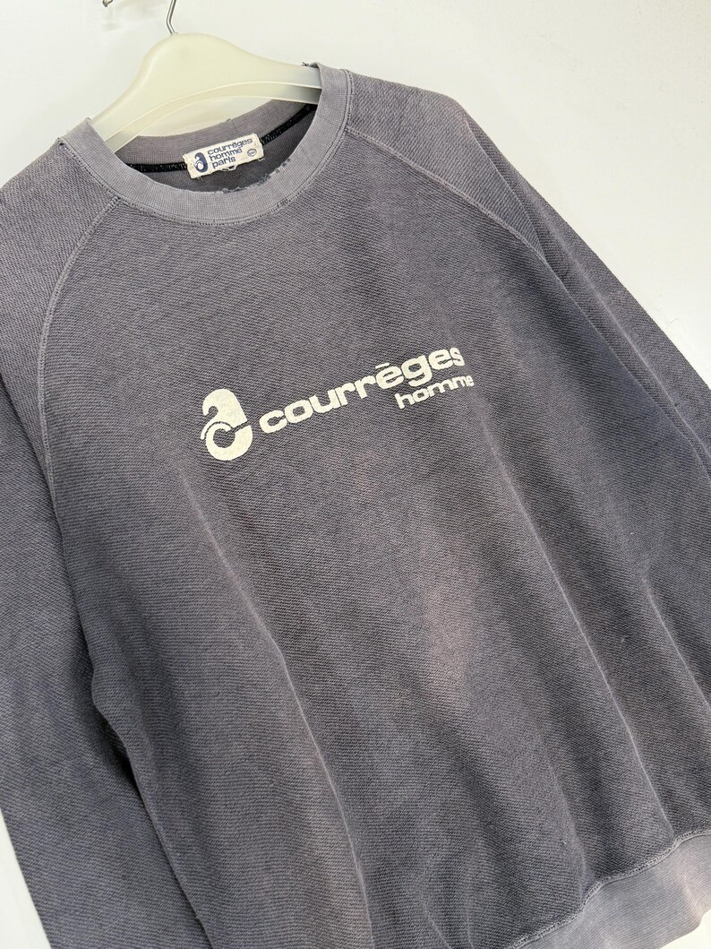 May include: A gray crew neck sweatshirt with the text "courr&egrave;ges homme" in white. The sweatshirt has a ribbed texture and a faded appearance. The brand label is visible at the neckline.