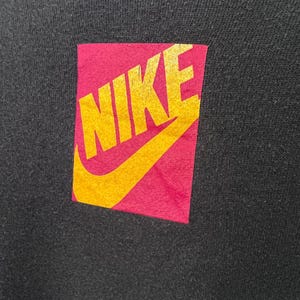 Puede incluir: Prenda negra con un parche rectangular con la palabra "NIKE" en dorado. El parche tiene un fondo rosa y un dise&ntilde;o de swoosh dorado.
