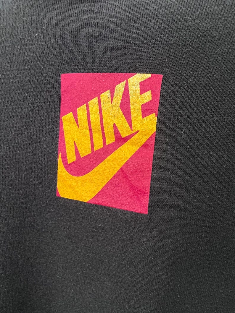 Puede incluir: Prenda negra con un parche rectangular con la palabra "NIKE" en dorado. El parche tiene un fondo rosa y un dise&ntilde;o de swoosh dorado.