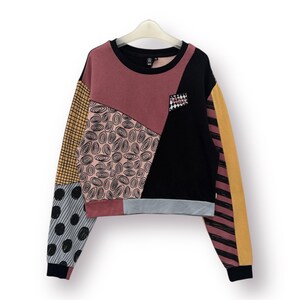 May include: A patchwork sweatshirt with a black crew neck and a small pocket. The design incorporates various patterns and colours, including black, pink, yellow, and grey. The sleeves have different patterns, and the bottom hem is cropped.