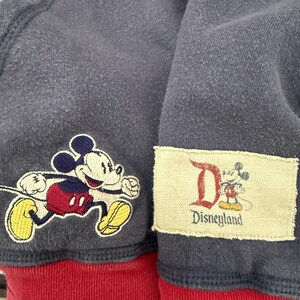 May include: Close-up of a navy blue zip-up hoodie with a red bottom hem. Features a running Mickey Mouse patch and a Disneyland patch. The hoodie is made of a soft, textured fabric.