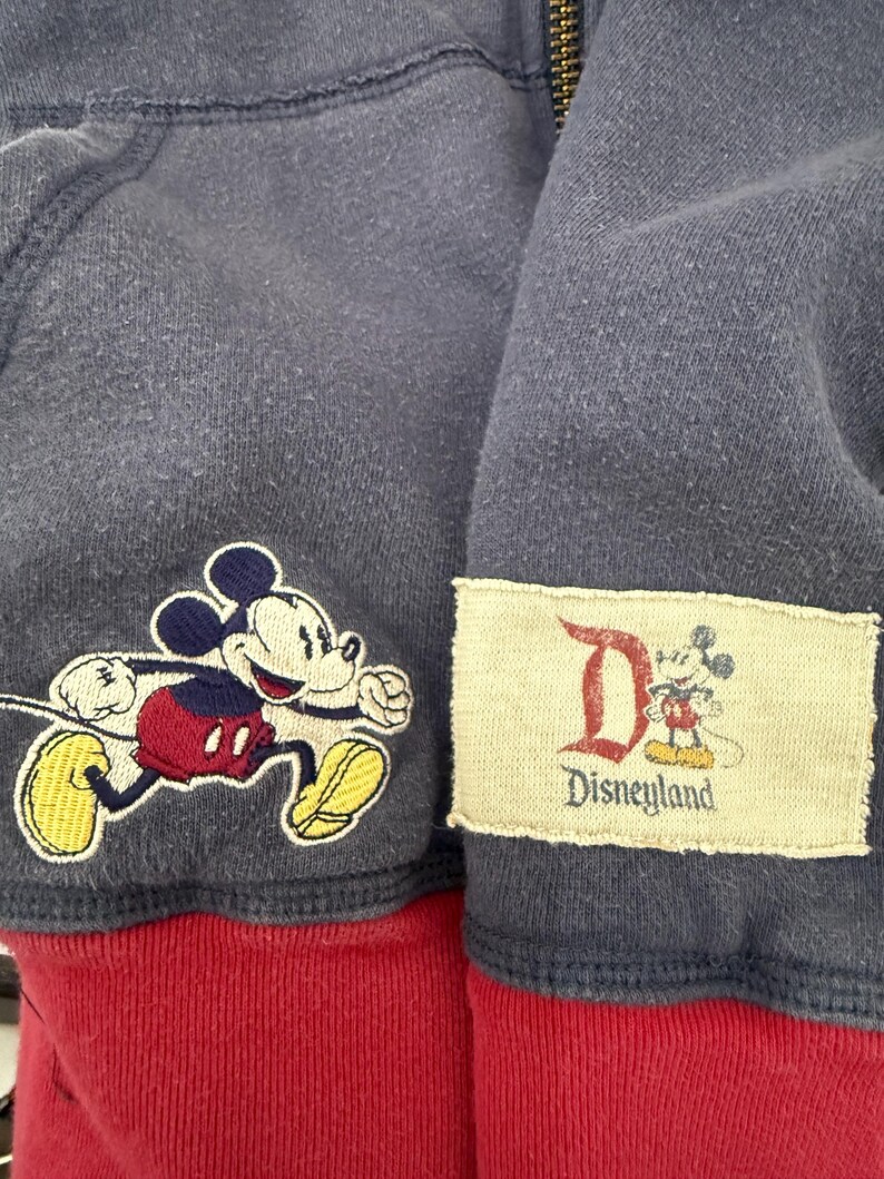 May include: Close-up of a navy blue zip-up hoodie with a red bottom hem. Features a running Mickey Mouse patch and a Disneyland patch. The hoodie is made of a soft, textured fabric.