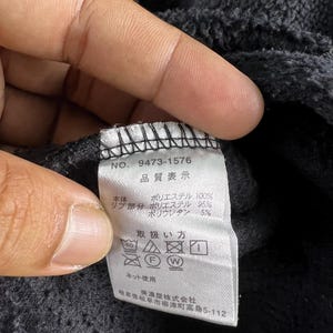 Op de afbeelding: Een zwart kledinglabel met witte tekst die "NO. 9473-1576", "品質表示", "本体 ポリエステル 100%", "リブ部分 ポリエステル 95%", "ポリウレタン 5%", "取扱い方", "ネット使用", "岐阜県岐阜市柳津町高路5-112", "※激型株式会社" luidt.