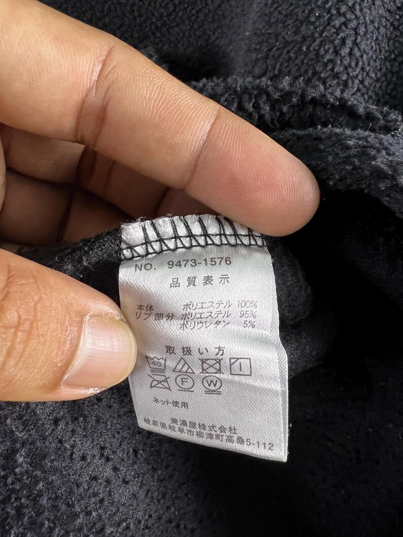 Op de afbeelding: Een zwart kledinglabel met witte tekst die "NO. 9473-1576", "品質表示", "本体 ポリエステル 100%", "リブ部分 ポリエステル 95%", "ポリウレタン 5%", "取扱い方", "ネット使用", "岐阜県岐阜市柳津町高路5-112", "※激型株式会社" luidt.