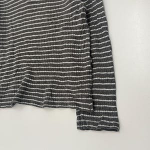 May include: A long-sleeve ribbed shirt featuring a dark gray and white horizontal striped pattern. The garment includes a quarter-zip neckline with a white zipper pull and a small square logo patch on the upper left chest area.