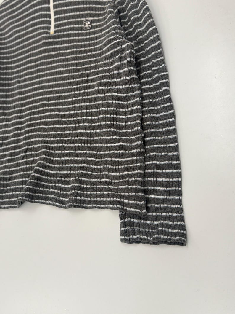 May include: A long-sleeve ribbed shirt featuring a dark gray and white horizontal striped pattern. The garment includes a quarter-zip neckline with a white zipper pull and a small square logo patch on the upper left chest area.