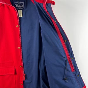 May include: Red jacket with a blue lining and a zipper closure. The jacket has a pocket on the front and a label that reads "THE WOOLRICH WOMAN".