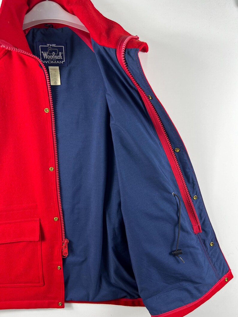 May include: Red jacket with a blue lining and a zipper closure. The jacket has a pocket on the front and a label that reads "THE WOOLRICH WOMAN".