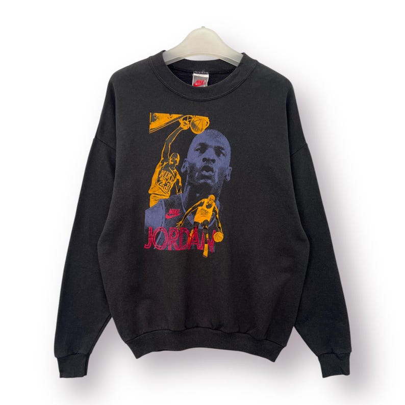 Puede incluir: Sudadera negra con un dise&ntilde;o gr&aacute;fico de un jugador de baloncesto en acci&oacute;n, con el nombre "JORDAN" en rojo. El dise&ntilde;o incluye im&aacute;genes en amarillo, morado y naranja. Un peque&ntilde;o logotipo de Nike es visible en el cuello.