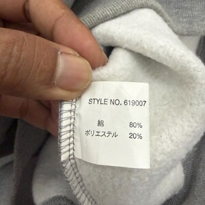 Pode incluir: Close-up do interior de um moletom cinza com uma etiqueta branca. A etiqueta exibe "STYLE NO. 619007" e a composi&ccedil;&atilde;o do tecido: 80% algod&atilde;o e 20% poli&eacute;ster. O moletom &eacute; segurado por uma m&atilde;o.