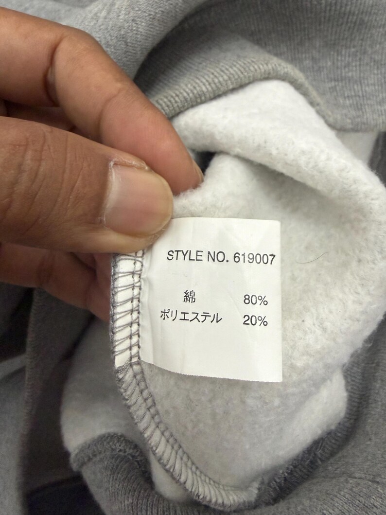 Pode incluir: Close-up do interior de um moletom cinza com uma etiqueta branca. A etiqueta exibe "STYLE NO. 619007" e a composi&ccedil;&atilde;o do tecido: 80% algod&atilde;o e 20% poli&eacute;ster. O moletom &eacute; segurado por uma m&atilde;o.