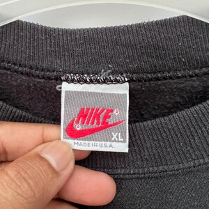 Puede incluir: Primer plano de una etiqueta de sudadera negra con el logo de Nike en rojo. La etiqueta dice "NIKE" y "XL" con "MADE IN U.S.A." impreso debajo. La sudadera es negra.