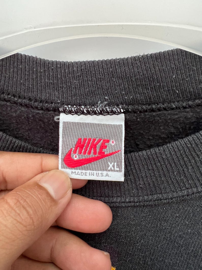 Puede incluir: Primer plano de una etiqueta de sudadera negra con el logo de Nike en rojo. La etiqueta dice "NIKE" y "XL" con "MADE IN U.S.A." impreso debajo. La sudadera es negra.