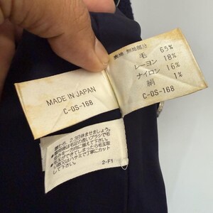 May include: Close-up of a navy blue garment with attached tags. One tag reads "MADE IN JAPAN" and "C-OS-168." The other tag lists fabric composition: 65% wool, 18% rayon, 16% nylon, and 1% silk. Additional Japanese text is also present.