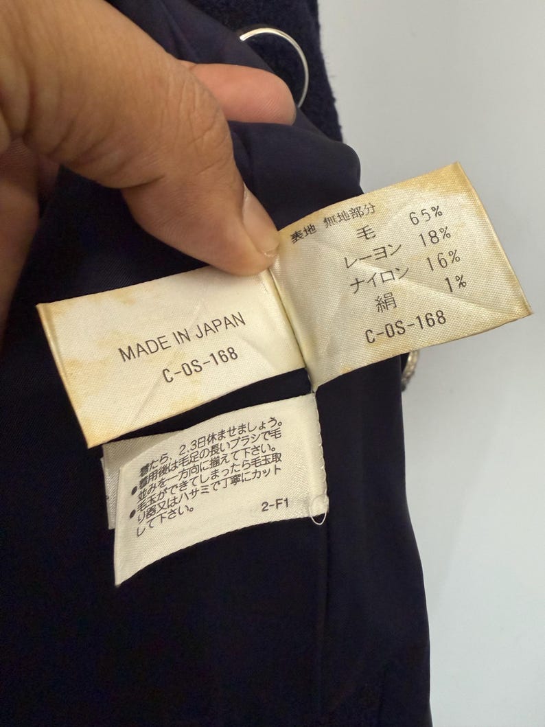 May include: Close-up of a navy blue garment with attached tags. One tag reads "MADE IN JAPAN" and "C-OS-168." The other tag lists fabric composition: 65% wool, 18% rayon, 16% nylon, and 1% silk. Additional Japanese text is also present.