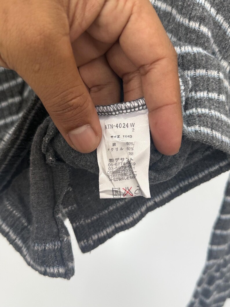 May include: A dark gray garment with light gray horizontal stripes and a white interior care label. The label displays the text ATN-4024 W, size 11(40), 50% cotton, 50% acrylic, and various Japanese characters including manufacturer contact information.