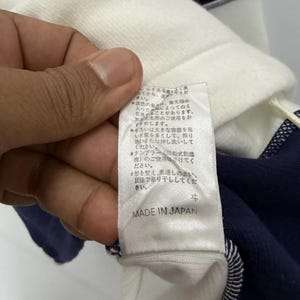 Puede incluir: Primer plano de una camisa a rayas blancas y azul marino. La camisa tiene una etiqueta que dice "MADE IN JAPAN". La etiqueta tambi&eacute;n tiene texto en japon&eacute;s. La camisa est&aacute; hecha de un material suave.