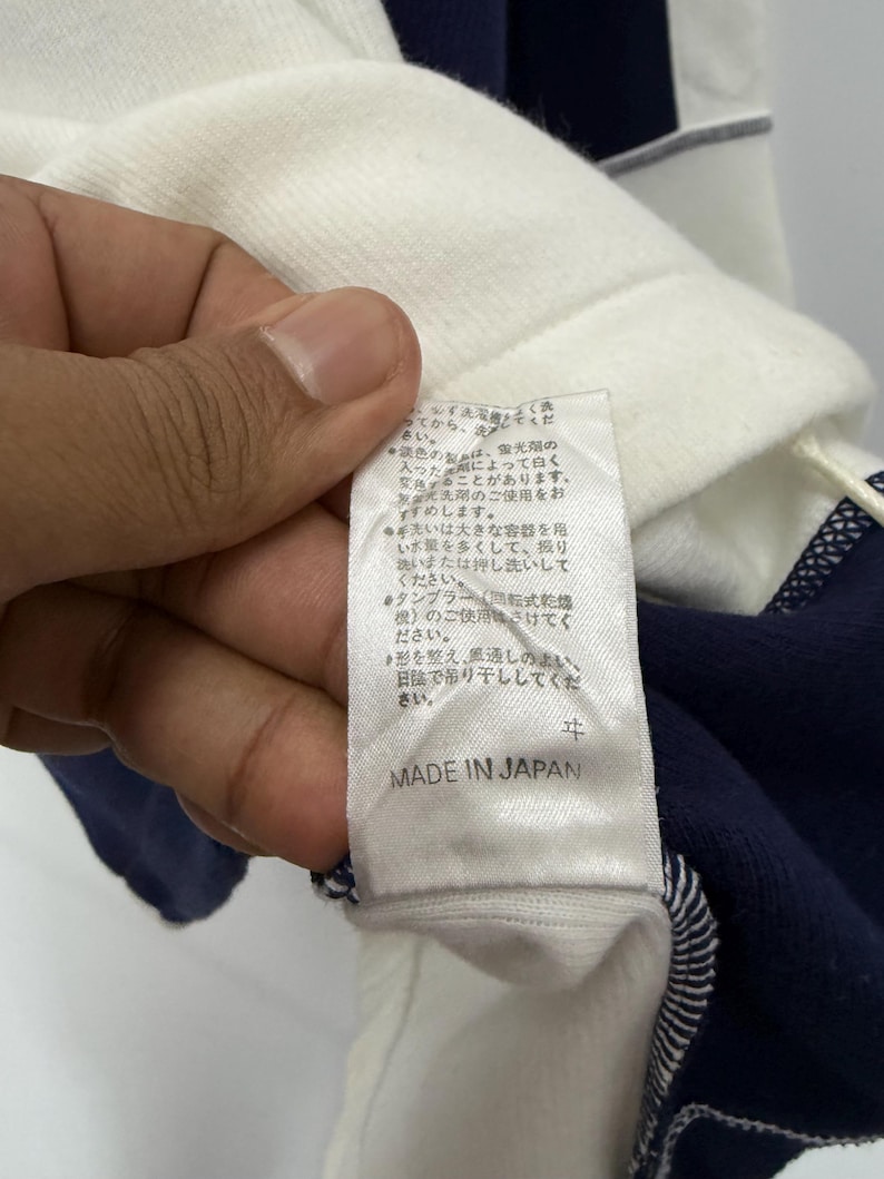 Puede incluir: Primer plano de una camisa a rayas blancas y azul marino. La camisa tiene una etiqueta que dice "MADE IN JAPAN". La etiqueta tambi&eacute;n tiene texto en japon&eacute;s. La camisa est&aacute; hecha de un material suave.
