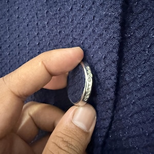 May include: A silver ring with the word "chuttes" engraved on the side. The ring is held against a textured navy blue fabric.