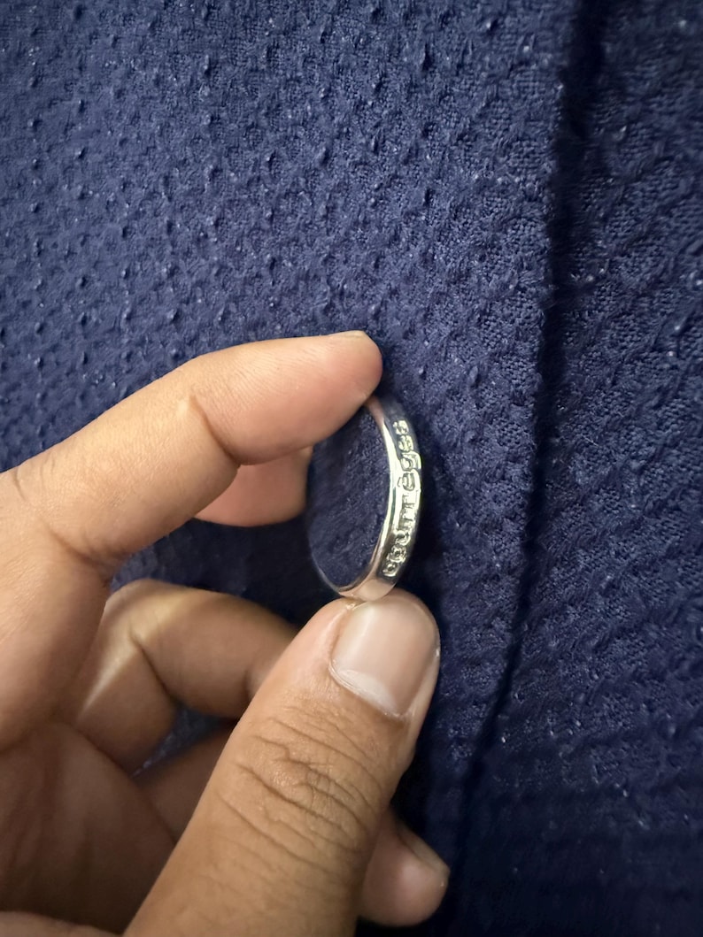 May include: A silver ring with the word "chuttes" engraved on the side. The ring is held against a textured navy blue fabric.