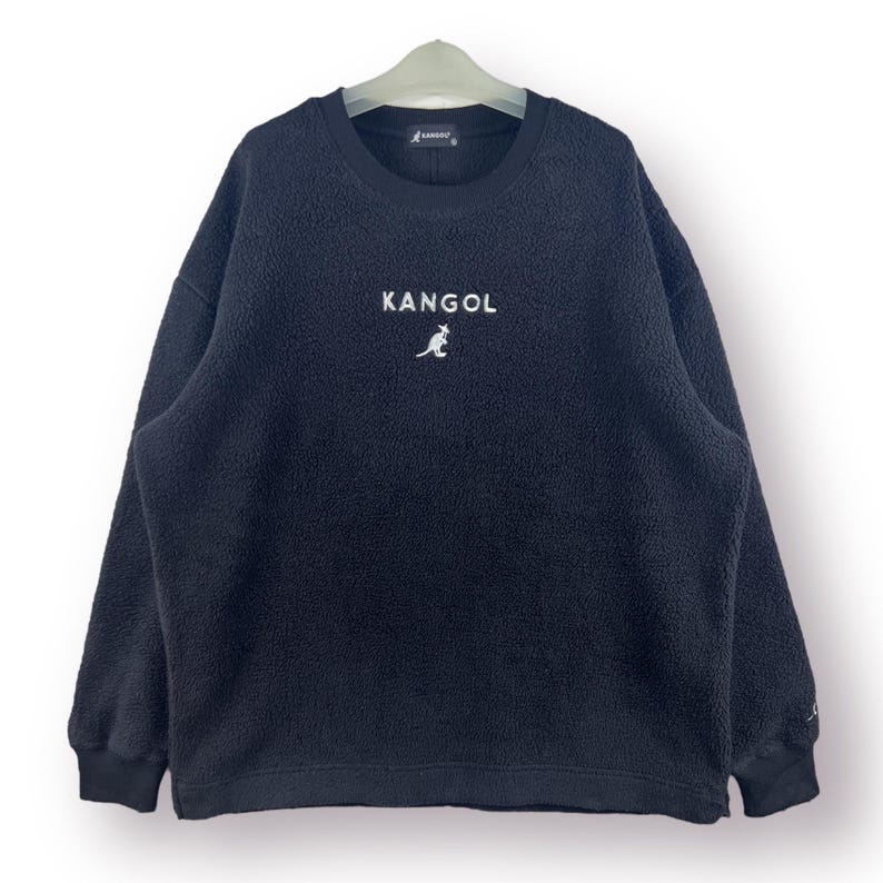 Op de afbeelding: Zwarte fleece trui met een wit geborduurd kangoeroe logo en het woord "Kangol" op de voorkant.