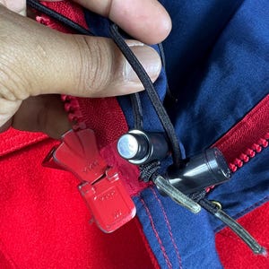 May include: Close-up of a red and blue jacket zipper with a YKK zipper pull. The zipper is partially open and the zipper pull is in the up position.