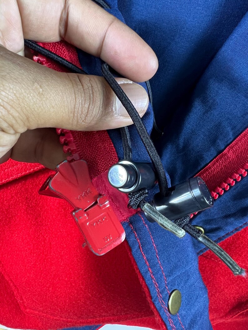 May include: Close-up of a red and blue jacket zipper with a YKK zipper pull. The zipper is partially open and the zipper pull is in the up position.
