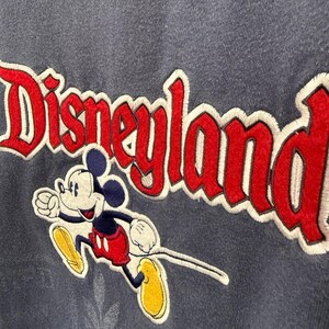 May include: A navy blue sweatshirt featuring the embroidered word "Disneyland" in red with a white outline. Mickey Mouse is depicted in a running pose, wearing red shorts, yellow shoes, and white gloves.