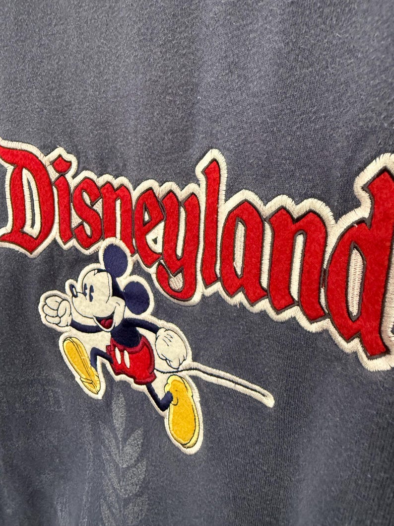 May include: A navy blue sweatshirt featuring the embroidered word "Disneyland" in red with a white outline. Mickey Mouse is depicted in a running pose, wearing red shorts, yellow shoes, and white gloves.