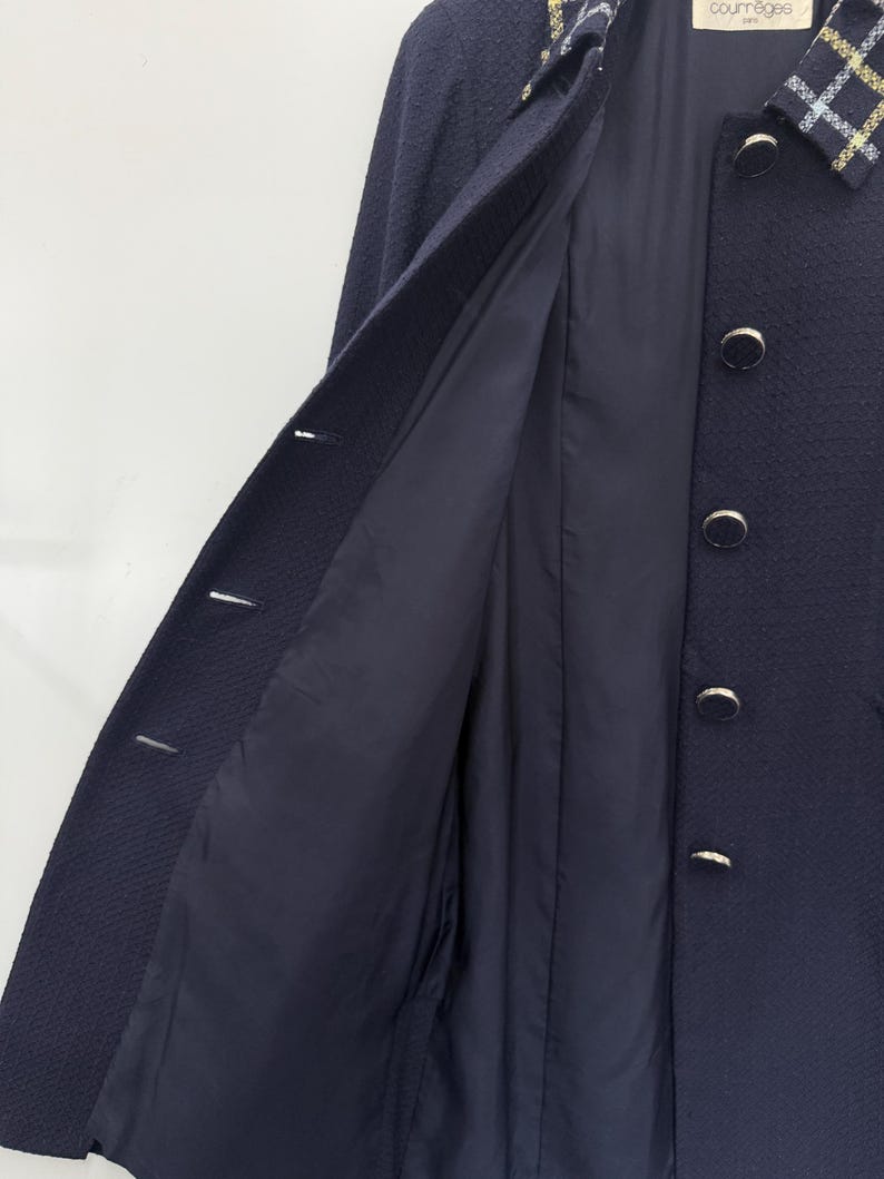 May include: A navy blue coat with a textured weave, featuring a contrasting plaid collar and gold-toned buttons. The coat's interior lining is a solid navy blue. The Courr&egrave;ges logo is visible on the collar.
