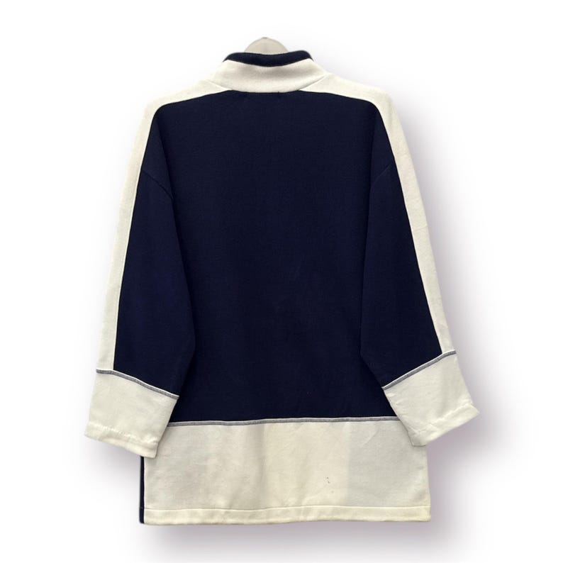 Puede incluir: Una camisa de manga larga azul marino y crema. La camisa presenta un cuello alto y paneles de color crema en las mangas y el dobladillo inferior. Las mangas tienen un ribete gris. La camisa est&aacute; hecha de un material suave.