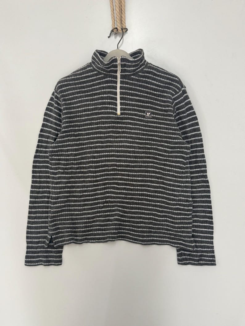 May include: A charcoal gray and white horizontally striped long-sleeve pullover sweater with a ribbed texture. The garment features a high collar with a white front zipper closure and a small square logo patch on the left chest area.