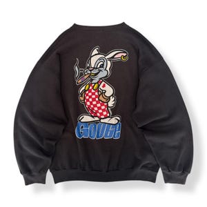 Könnte beinhalten: Schwarzes Sweatshirt mit einem Cartoon-Hasen, der eine Zigarre raucht, eine rot-weiß karierte Schürze und einen goldenen Ohrring trägt. Das Wort "GOUGE" ist unten in Blau aufgedruckt. Der Hase steht auf einer blauen Plattform.