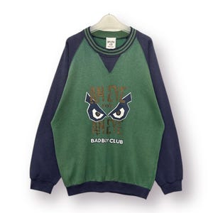 Pode incluir: Uma camisola verde e azul marinho com o texto "AN EYE FOR AN EYE" e "BAD BOY CLUB" estampado na frente. A camisola tem gola redonda e mangas compridas.