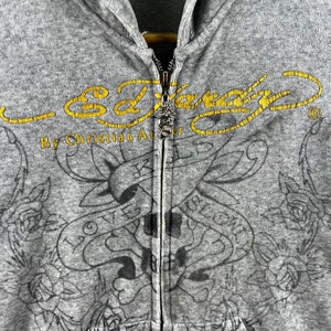 May include: Gray hooded sweatshirt with a black and white tattoo design and the text "Ed Hardy By Christian Audigier" in gold lettering.
