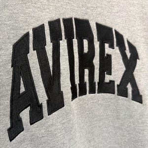 Pode incluir: Close-up de um moletom cinza com a palavra "AVIREX" em letras pretas arqueadas. As letras grossas e texturizadas contrastam com o tecido liso do moletom, criando um efeito visual.