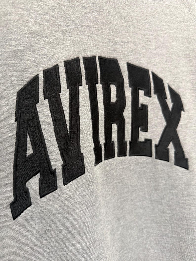 Pode incluir: Close-up de um moletom cinza com a palavra "AVIREX" em letras pretas arqueadas. As letras grossas e texturizadas contrastam com o tecido liso do moletom, criando um efeito visual.