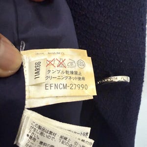 May include: Close-up of a navy blue garment with a tag featuring Japanese text and care symbols. The tag includes the code "11AR66" and "EFNCM-27990". The garment has a silver-colored clasp.